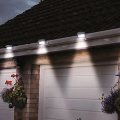 three lights on the side of a building near flowers and potted plants in front of a garage door