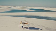 two people sitting at a white piano in the middle of an expanse of sand and water