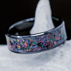 a black ring with multicolored opal inlays on it sitting on top of a rock