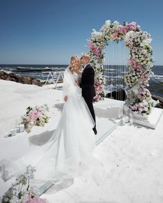 a bride and groom standing in front of an arch with flowers on it near the ocean