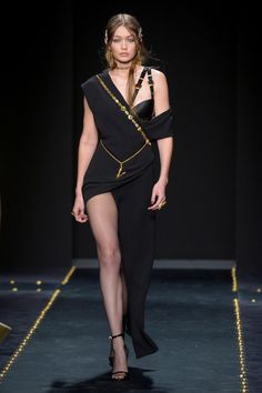 a model walks down the runway in a black dress with gold chains around her neck