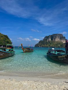 three boats are docked on the beach near some mountains and clear blue water with people standing in the water