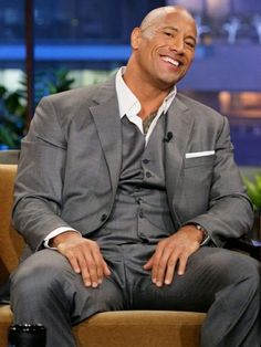 a smiling man in a gray suit and white shirt sitting on a chair with his legs crossed