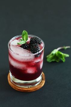 a blackberry cocktail with ice and mint garnish on a black surface next to leaves