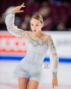 a female figure skating on the ice in a white dress with an open shoulder and long sleeves