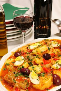 a bowl of food with olives, potatoes and other foods on the table next to wine glasses