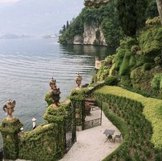 an outdoor area with hedges and benches on the side of a body of water, surrounded by greenery