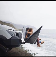 two people sitting in a hammock next to the ocean on a cloudy day