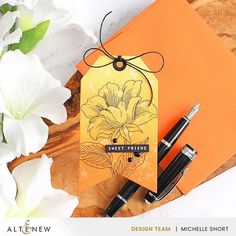 an orange card with flowers and two pens sitting on top of it next to some white flowers