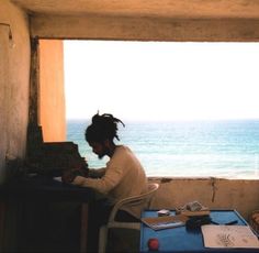 a woman sitting at a table in front of an open window looking out to the ocean