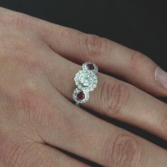 Edwardian Three Stone Moissanite Ruby Flower Halo Diamond Forever One Floral Engagement Ring Ornate and romantic three stone engagement ring made of 14K white gold. Ring features two round, bezel set red rubies (3.5mm each, .38CT) on either side of a round, 5mm (~.5ct) Forever One Moissanite center stone. All three stones are surrounded by flower shape halos of pave-set, round diamonds (.36cttw, G-VS quality). High polish, smooth sides and edges, cut-out baskets support bigger stones. Dimensions Anniversary Ruby Ring With Moissanite In Round Cut, Anniversary Ruby Ring With Round Cut Moissanite, White Ruby Ring With Brilliant Cut, Wedding Ruby Ring With Brilliant Cut In Diamond White, Brilliant Cut Ruby Ring In Platinum For Anniversary, Formal White Gold Ruby Ring With Three Stones, Heirloom Three Stone Ruby And Diamond Ring, Diamond White Ruby Ring With Center Stone, White Gold Rose Cut Diamond Proposal Ring