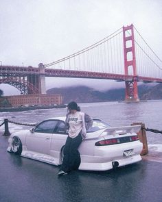 a person leaning on the hood of a car in front of a bridge and water