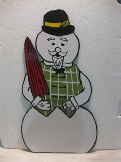 a white snowman holding a red ski in his hand and wearing a green plaid shirt