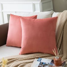 two pink pillows sitting on top of a white couch next to a book and plant