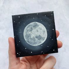 a hand holding up a small square painting with the moon in it's center