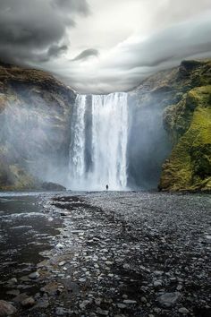 a man standing in the middle of a river next to a tall waterfall under a cloudy sky