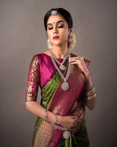 a woman in a green and pink saree with jewelry on her neck, standing against a gray background