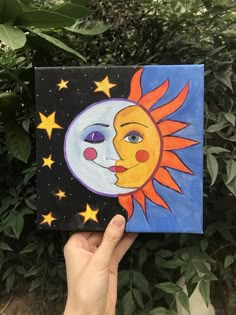 a hand holding up a small painting with the sun and moon on it in front of some bushes