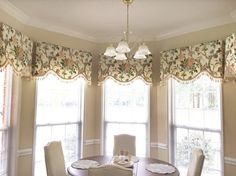 a dinning room table with four chairs in front of three windows covered by valances