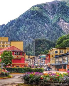 a town with mountains in the background and colorful flowers growing on the sidewalk, surrounded by greenery