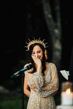 a woman wearing a crown standing in front of a microphone and holding her hands to her face