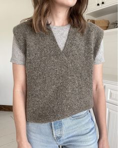 a woman standing in a kitchen wearing a sweater vest and jeans with her hands in her pockets