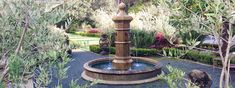 an outdoor fountain surrounded by trees and plants in a garden area with a cat sitting on the ground next to it