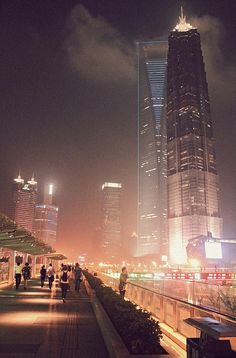 people are walking on the sidewalk in front of tall buildings at night time with lights shining