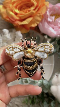 a hand holding a bee brooch in front of flowers