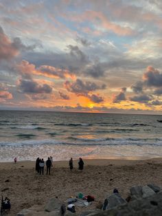 people are standing on the beach watching the sun go down over the water and clouds