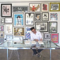 a man sitting at a glass desk in front of a wall full of framed posters