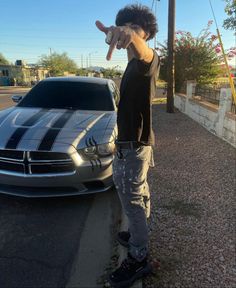 a young man pointing at the camera next to a silver sports car in a parking lot