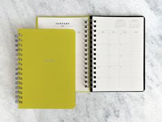 a yellow and black spiral bound notebook next to a calendar on a white marble surface