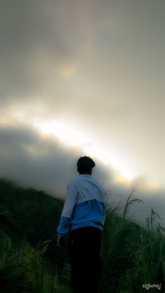 a man standing on top of a lush green hillside under a cloudy sky in the distance