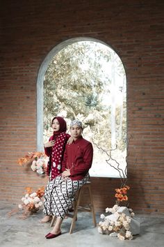 two people sitting on a chair in front of an arched window with flowers around them