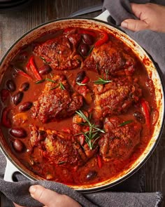 a pan filled with meat and olives on top of a wooden table next to a person's hand