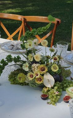 an arrangement of flowers, fruit and greenery is displayed on a table in front of a chair