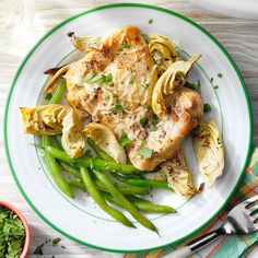 a white plate topped with chicken and artichokes next to a bowl of green beans