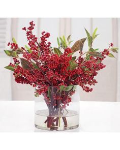 a glass vase filled with red berries and greenery on top of a white table