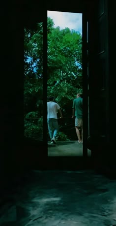 two people walking through an open door in the dark, with trees behind them and one person wearing white shirt