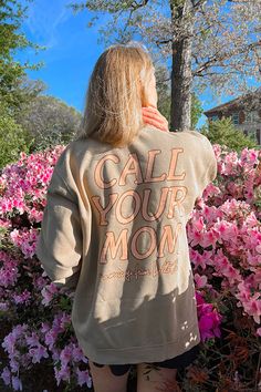 a woman standing in front of pink flowers with her back to the camera, wearing a sweatshirt that says call your mom