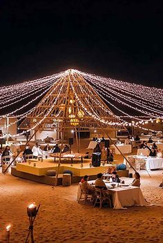 people are sitting at tables in the sand under a lit up tent with lights on it