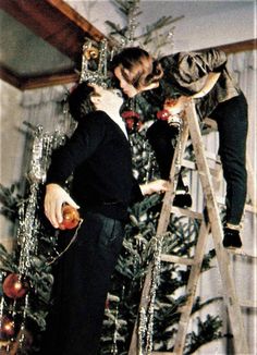 two people standing on a ladder next to a christmas tree and one is kissing the other