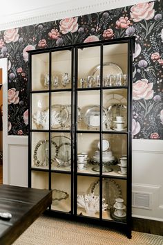 a china cabinet with glass doors and floral wallpaper on the walls in a dining room