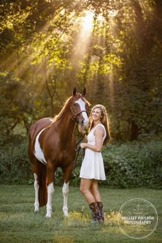 a beautiful young woman standing next to a brown and white horse in front of trees