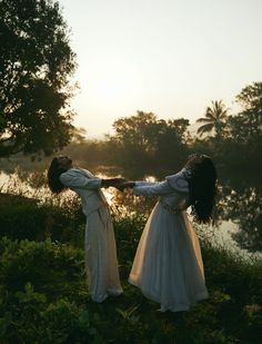 two women in white dresses are holding hands near the water at sunset or dawn,