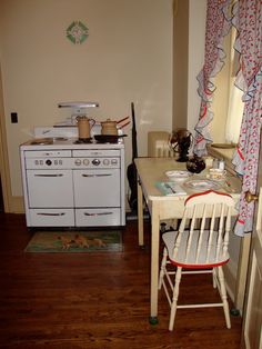 an old fashioned stove and table in a small room with wood floors, white cabinets and curtains