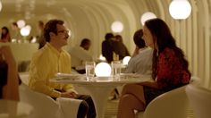 two people sitting at a table in a room with round lights on the ceiling and white chairs