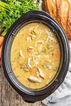 a crock pot filled with chicken soup next to bread and carrots on a wooden table