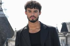 a man with curly hair wearing a suit and black shirt standing in front of an old building